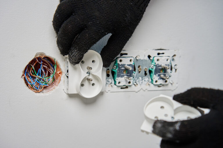 Close-up of an electrician wearing black gloves installing power socket on white wall. Electrical repair and maintenance process in progress