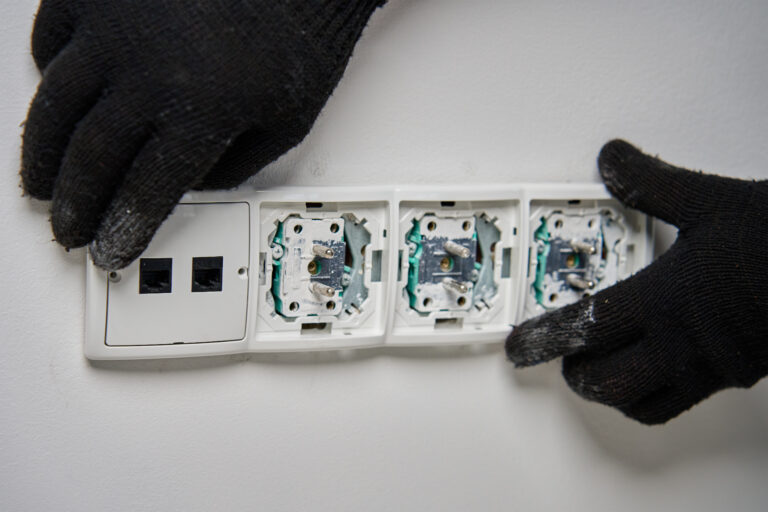 Close-up of an electrician wearing black gloves installing power socket on white wall. Electrical repair and maintenance process in progress